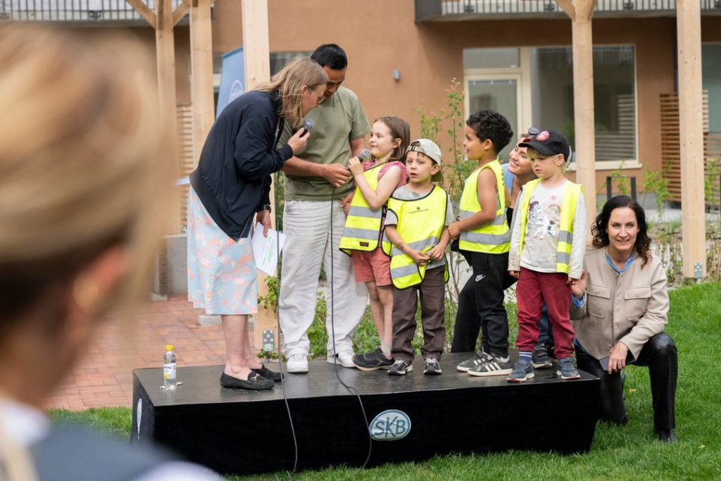 En grupp barn i reflexvästar står på en liten scen utomhus tillsammans med vuxna. En kvinna håller en mikrofon framför ett av barnen, medan en annan vuxen står bredvid. En kvinna sitter på scenkanten. I förgrunden syns en suddig person.