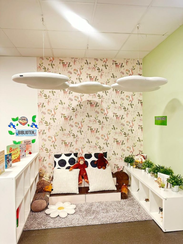 Mysig läshörna på förskola med en tapet mönstrad av djur som rådjur och rävar. En liten soffa fylld med kuddar och gosedjur, som en apa och en igelkott, står i mitten omgiven av vita bokhyllor med barnböcker och krukväxter. Molnformade lampor hänger i taket, och en skylt med texten 'BIBLIOTEK' syns till vänster.