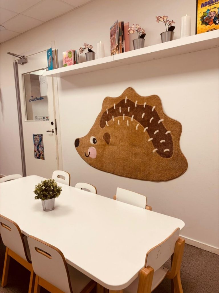Ljust rum på förskola med ett vitt bord och stolar, en dekorativ igelkottsformad matta på väggen, samt en hylla med böcker, ljus och krukväxter. En dörr med texten 'Junibacken' syns till vänster.