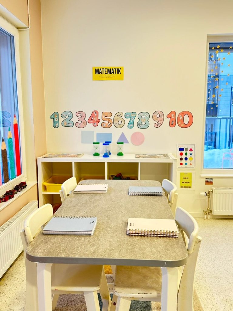 Matematikhörna på förskola med siffrorna 1 till 10 uppsatta på väggen ovanför en hylla med timglas och geometriska former. Ett bord med fyra stolar och anteckningsblock står i förgrunden.
