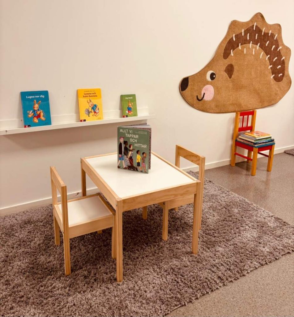 Läshörna på förskola med ett litet trä bord och stolar, en bokhylla med barnböcker samt en dekorativ igelkottsmatta på väggen. En röd stol med en trave böcker står bredvid mattan.