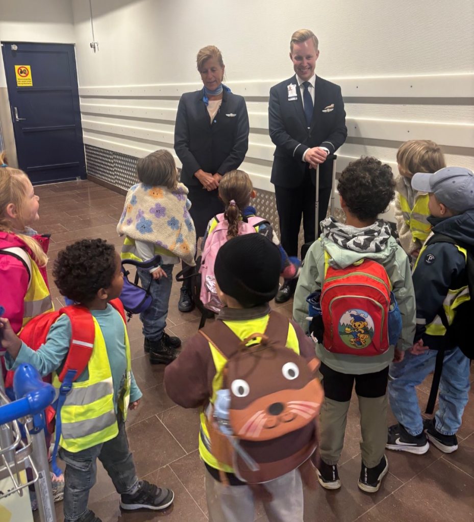 Grupp förskolebarn med ryggsäckar och reflexvästar lyssnar nyfiket på en pilot och en kabinanställd i uniform, troligtvis under ett studiebesök på en flygplats.