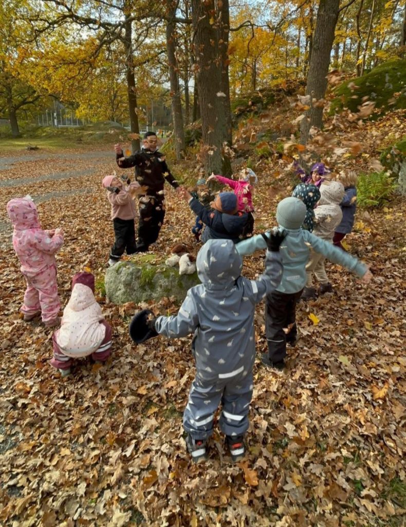 Förskolebarn i regnkläder kastar glatt höstlöv i luften tillsammans med en vuxen, i en skog full av gula och bruna löv