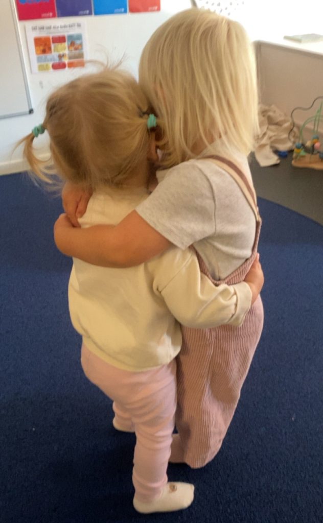 Två små barn kramar varandra ömt på en blå matta i ett rum på förskolan, med böcker och en anslagstavla synliga i bakgrunden.