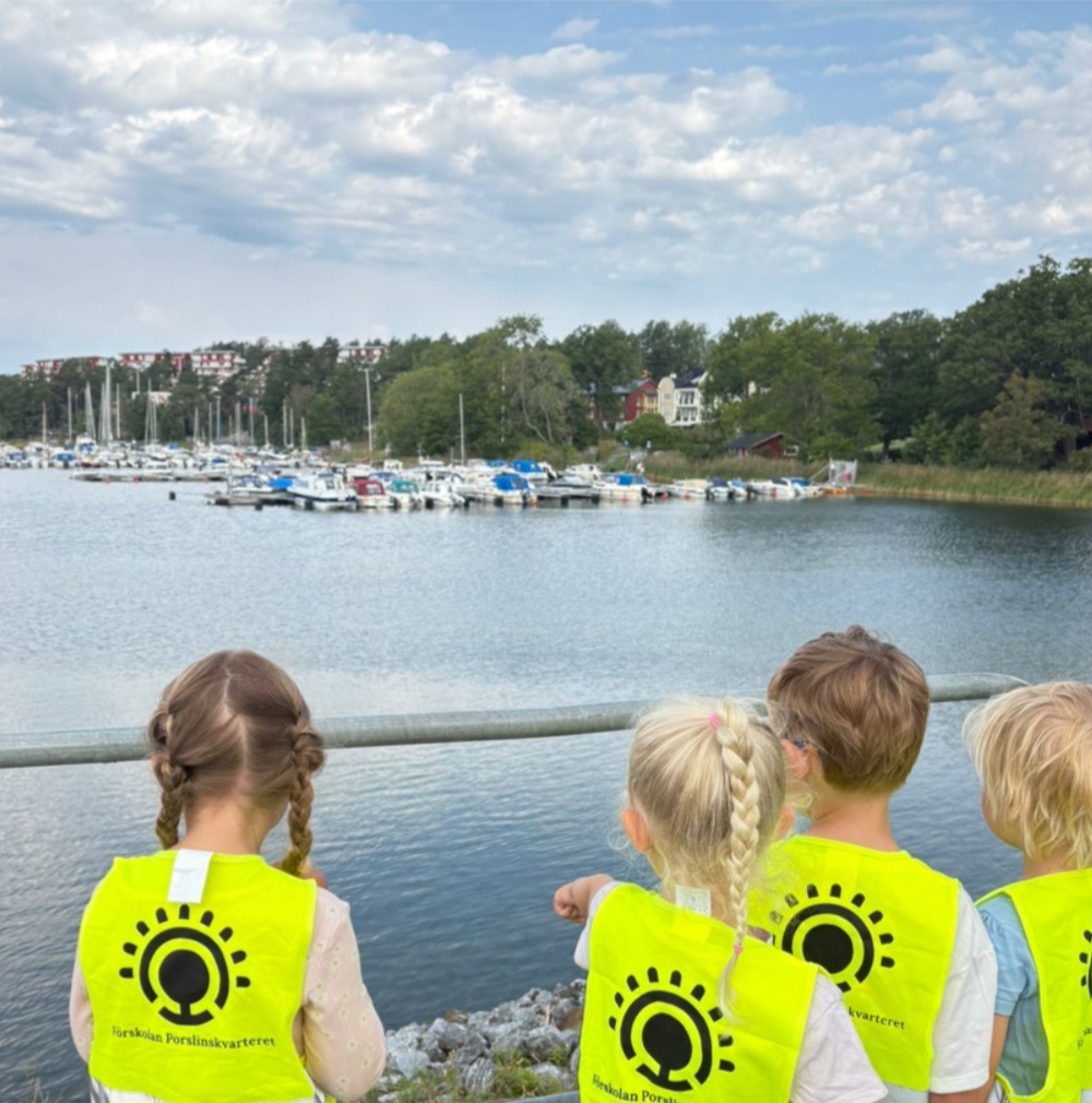 Fyra barn i gula reflexvästar med texten 'Förskolan Porslinskvarteret' står vid ett räcke och blickar ut mot en småbåtshamn med flera båtar, omgiven av grönska och bostadshus under en molnig himmel.