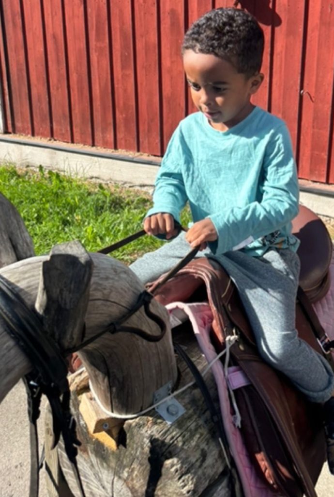 Ett barn sitter och håller i tyglarna på en trähäst med sadel, framför en röd träfasad.