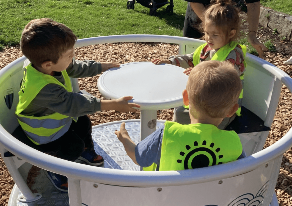 Tre barn i gula reflexvästar sitter runt ett vitt runt bord i en lekställning utomhus, med gräs och grusmark i bakgrunden.