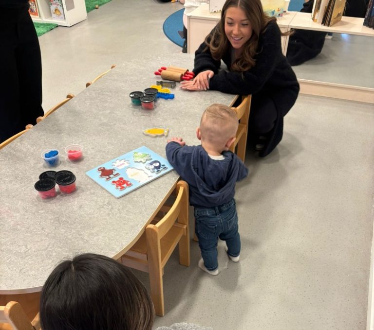 Ett barn sträcker sig över ett bord där ett halvfärdigt puzzel, ligger. En kvinna sitter på huk mittemot barnet och ler