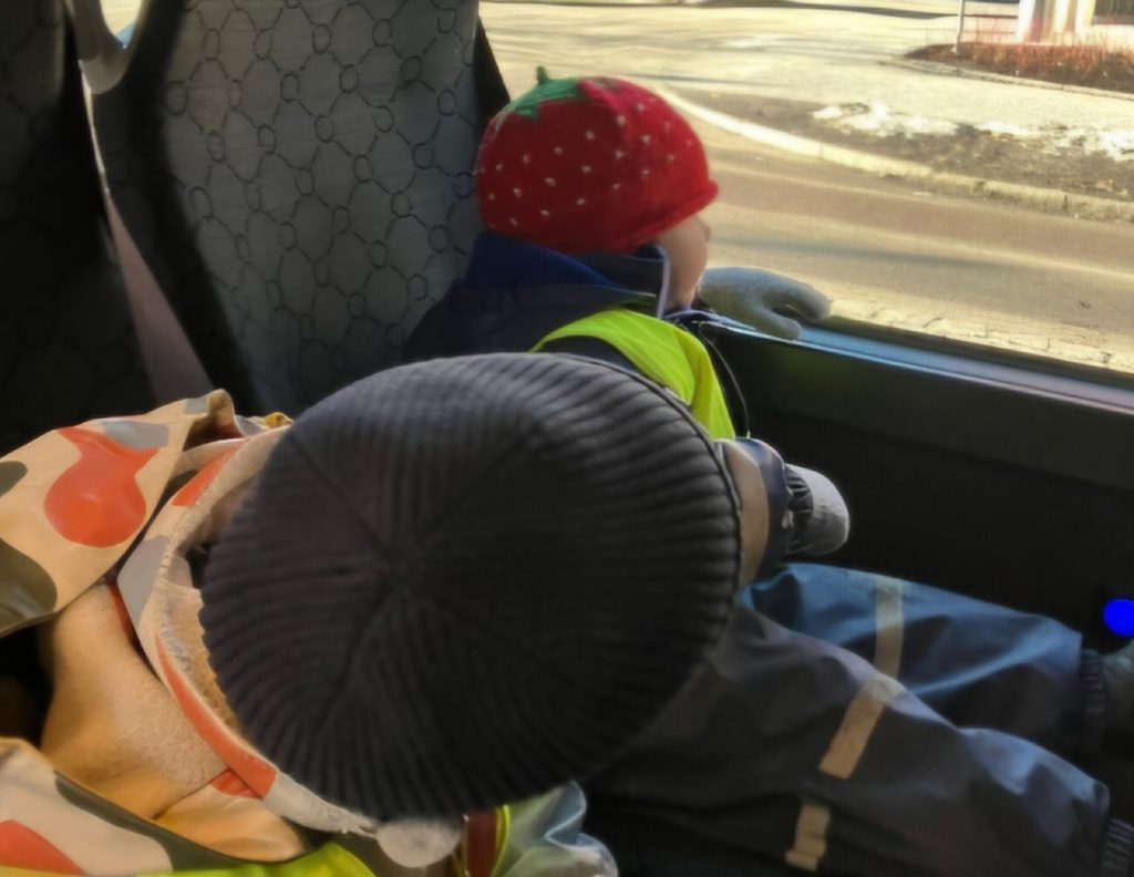 Två barn sitter i sittvagnar/bussäte och tittar ut genom fönstret, det ena i en röd jordgubbsmönstrad mössa och gul reflexväst.