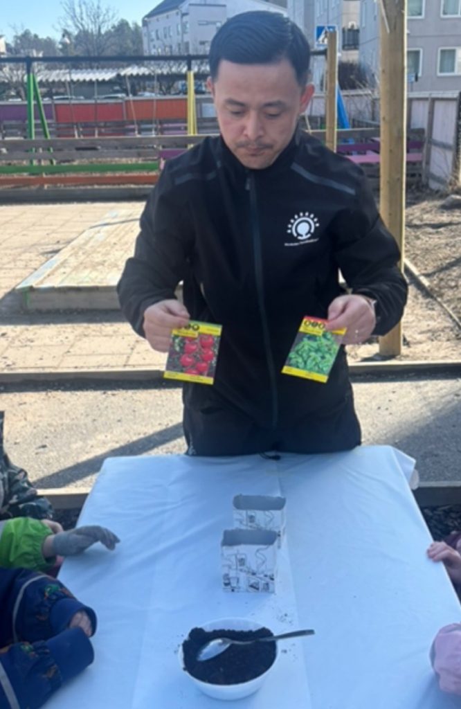 En vuxen i svart jacka håller upp fröpaket med tomat och basilika vid ett bord med jord och en frökruka, medan barn syns delvis i förgrunden utomhus.