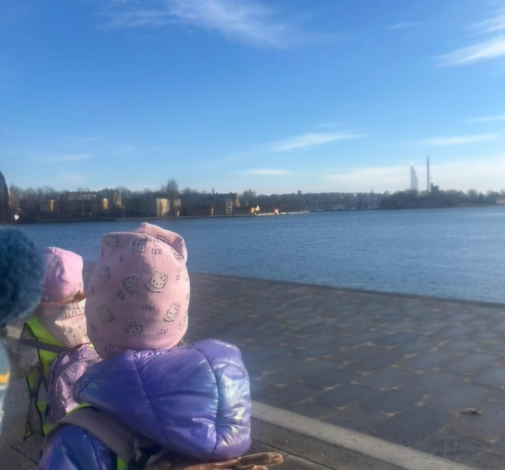Barn sitter vid en hamnkaj och blickar ut över vattnet mot en stadssiluett, under klarblå himmel.