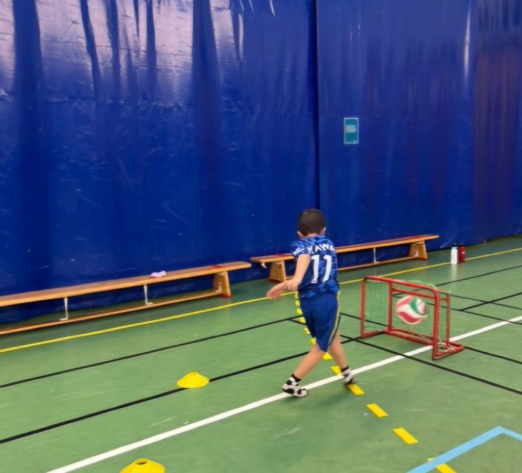 Ett barn i blå fotbollströja med nummer 11 sparkar en boll mot ett litet mål i en idrottshall, med koner på golvet.