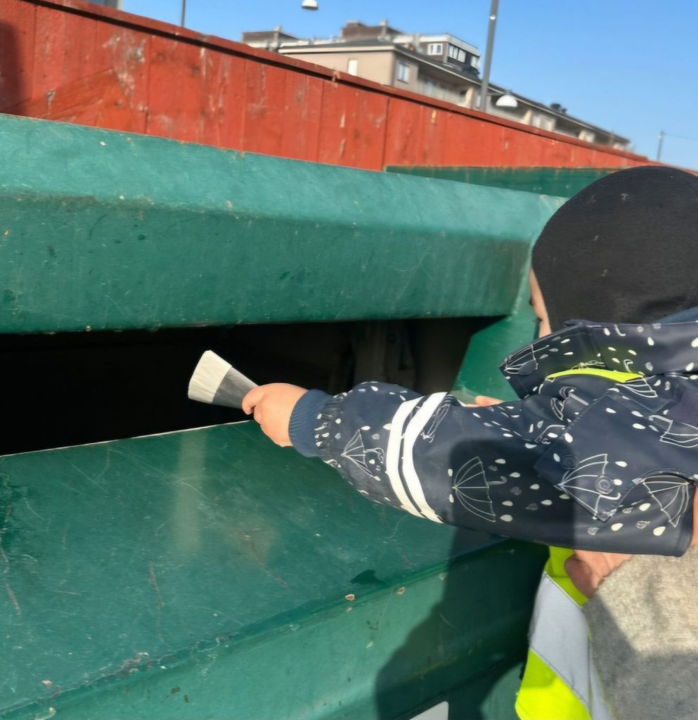 Ett barn i mörkblå prickig jacka och reflexväst sträcker sig med en borste mot en öppning i en stor grön container.