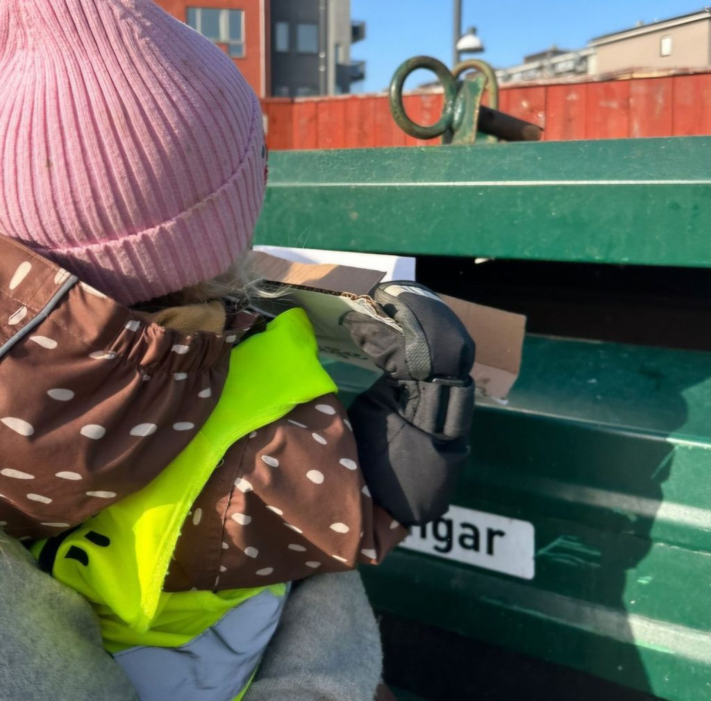 Närbild på ett barn i reflexväst och prickig jacka som lägger papper i en grön återvinningscontainer.
