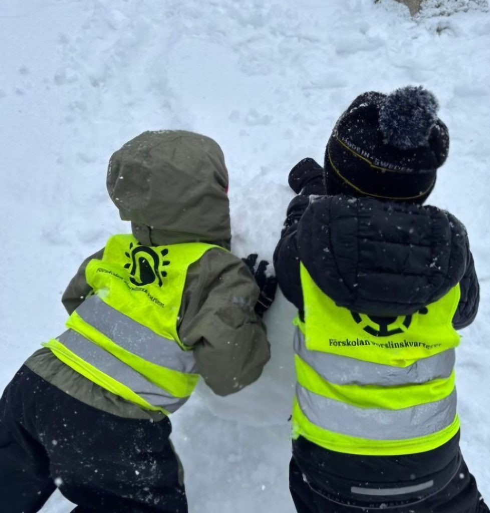 Två barn i gula reflexvästar från förskolan Porslinskvarteret bygger tillsammans i snön, sedda bakifrån.