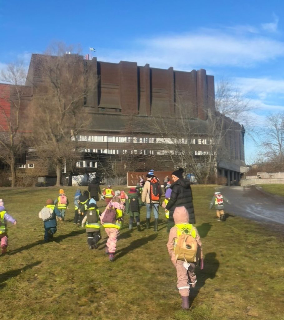 En grupp förskolebarn i gula reflexvästar går tillsammans över en gräsmatta mot en stor brun byggnad under klarblå himmel.