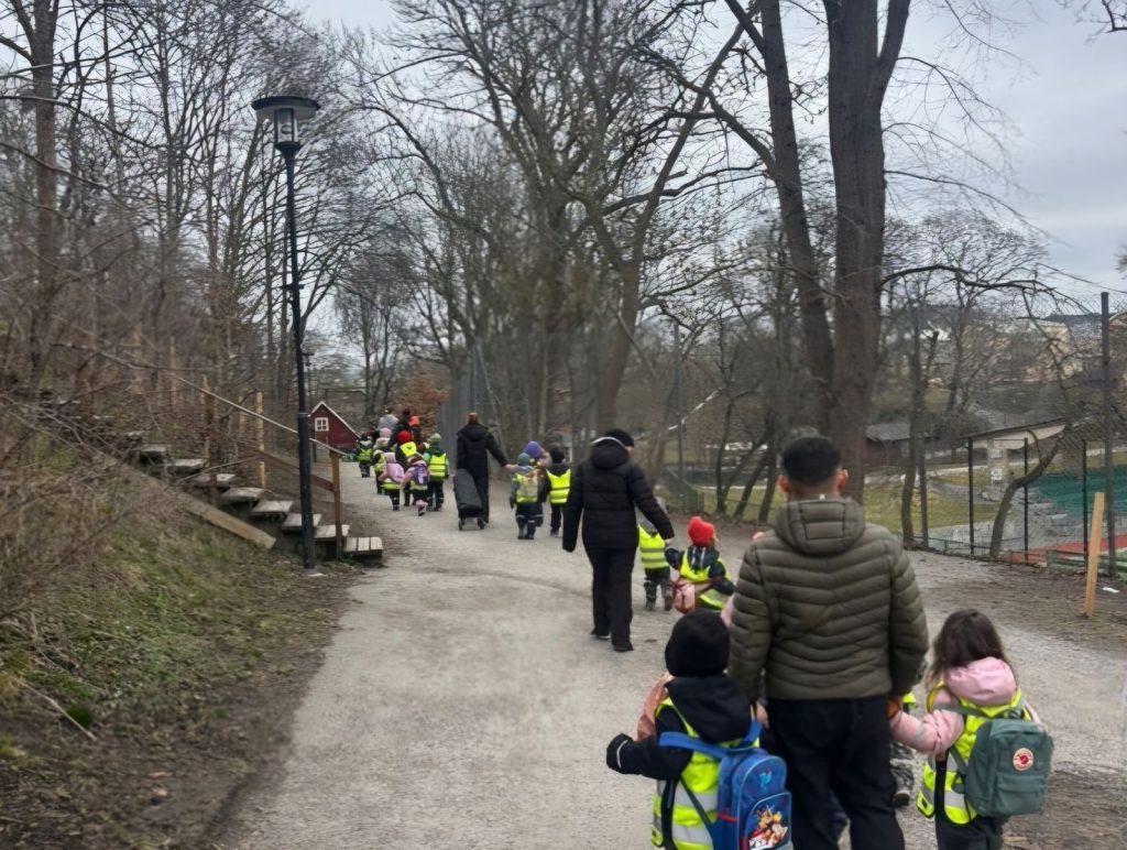 En grupp förskolebarn i gula reflexvästar och vuxna går tillsammans längs en grusväg omgiven av kala träd, med utsikt över ett vattendrag i bakgrunden.