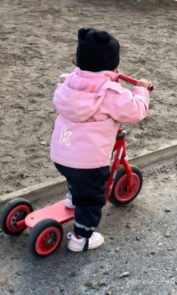 Ett barn i rosa jacka och svart mössa står på en sparkcykel och håller i styret på en röd balanscykel, på en sandig lekplats.