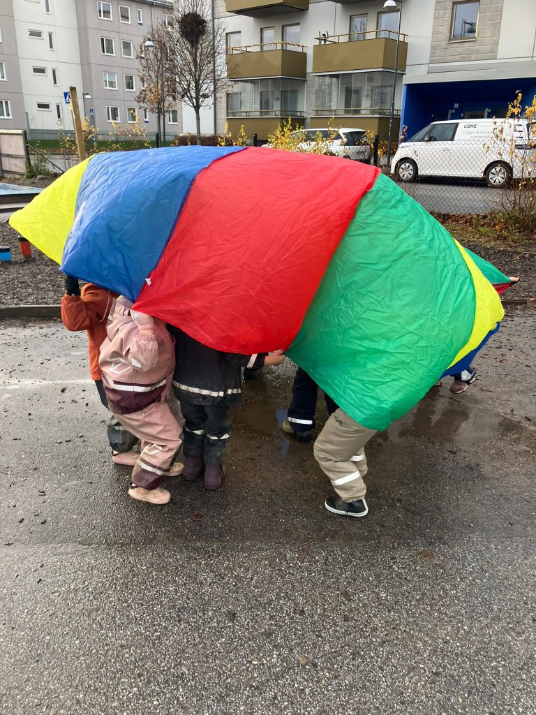 Barn står tillsammans under en stor regnbågsfärgad fallskärmsduk (blå, röd, gul, grön) utomhus på en asfalterad gård.