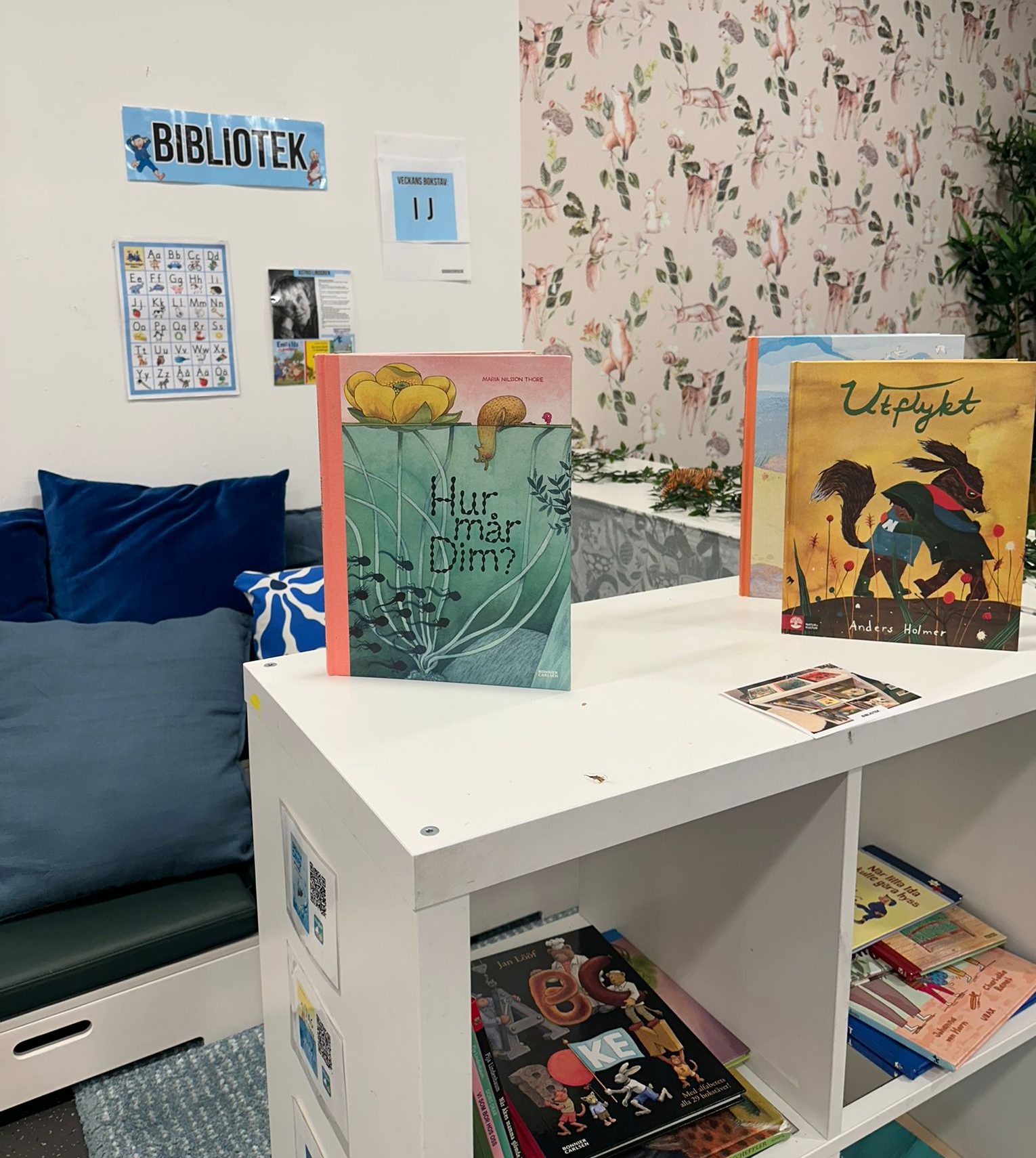 Bibliotekshörna på förskola med en vit bokhylla där barnböcker som 'Hur mår Dim?' och 'Utflykt' visas upp stående. På väggen sitter en skylt med texten 'BIBLIOTEK' samt ett alfabetsposter. Bakgrunden har en tapet med mönster av djur som rådjur och rävar, och till vänster syns en blå soffa med kuddar.