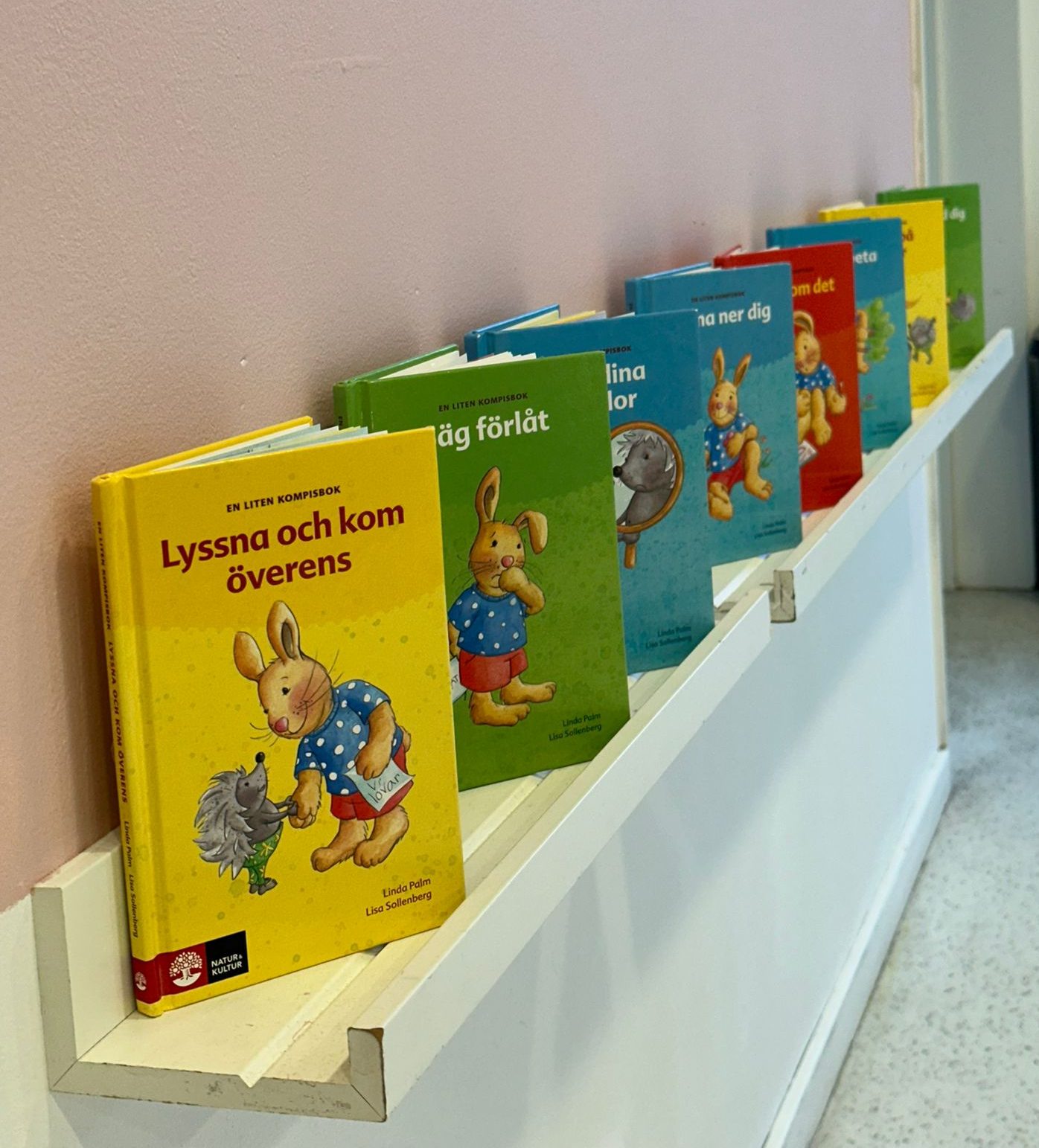 En rad böcker ur bokserien 'En liten kompisbok' med kanin- och igelkottsmotiv, uppställda på en vit vägghylla i en korridor. Böckerna har olika färgglada omslag i gult, grönt, blått och rött.