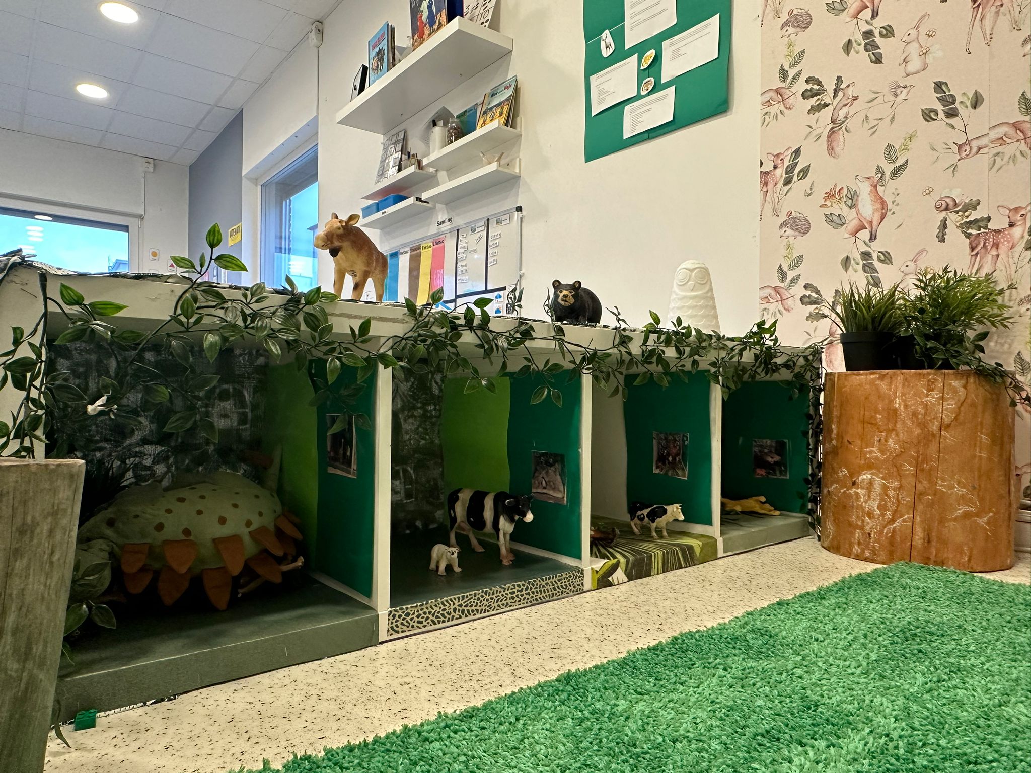 Skogstematiskt lekhörn på förskola med öppna hyllfack dekorerade som djurbon, gröna väggar och konstgjord murgröna. Leksaksdjur som kor, en björn och en älg är utplacerade, och en grön konstgräsmatta ligger på golvet framför