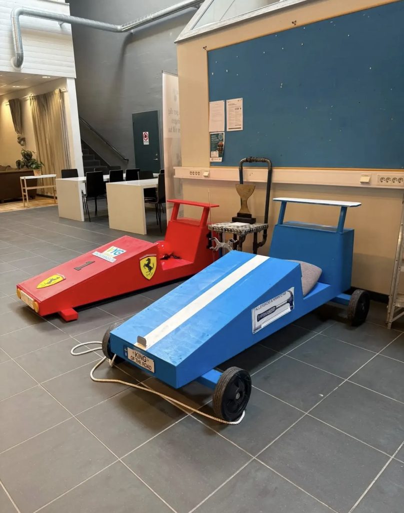 Två handbyggda sepetbilar (kanonlådebilar) i trä, en röd med Ferrari-emblem och en blå med texten "King of the Road", uppställda inomhus bredvid en pokal och en anslagstavla.