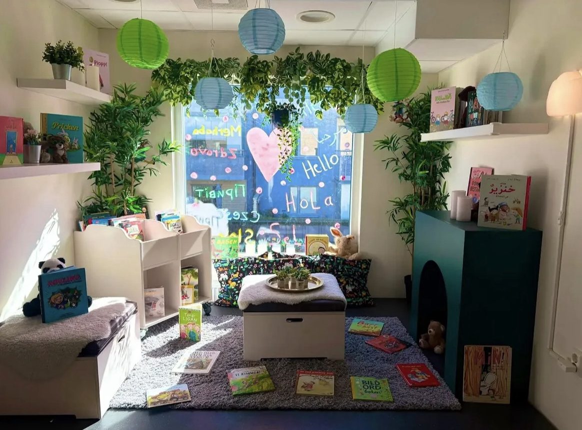 Mysig läshörna på förskola med gröna och blå papperslyktor hängande i taket, bokhyllor med barnböcker, en grå matta med utspridda böcker och gosedjur. Fönstret är dekorerat med ordet 'Hej' på olika språk och ett rosa hjärta, omgivet av grönskande krukväxter.