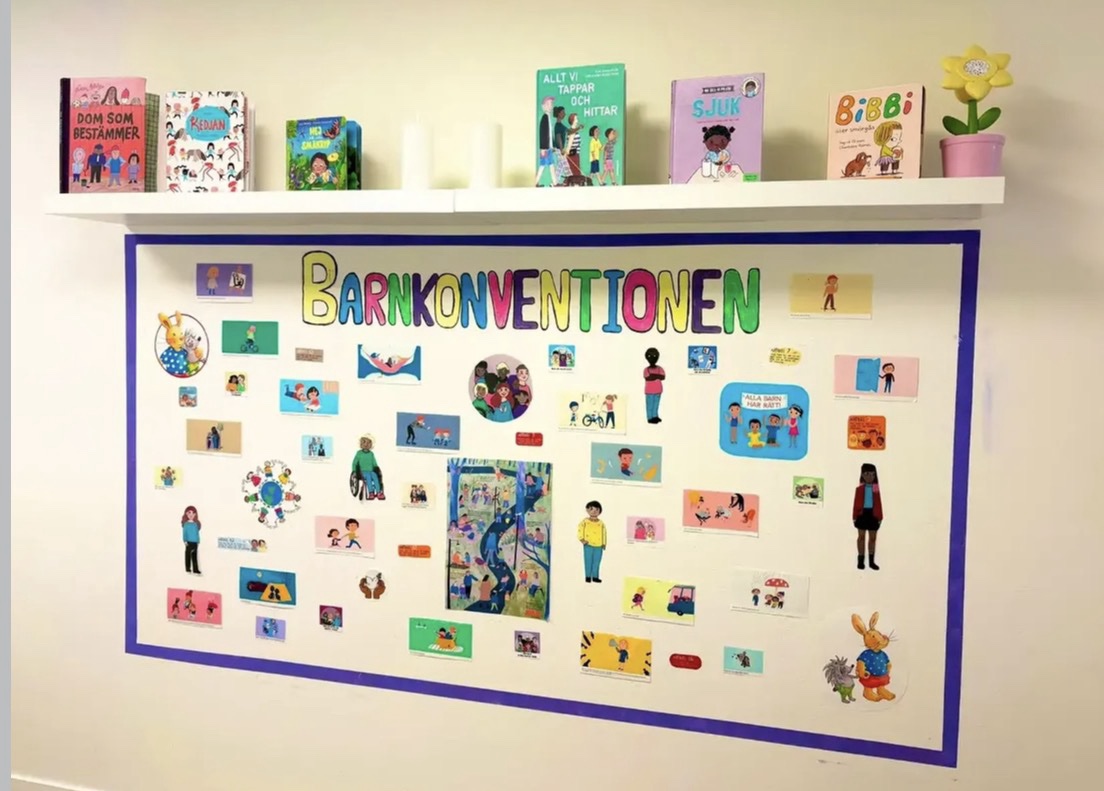 Anslagstavla på förskola med texten 'Barnkonventionen' i färgglada bokstäver, dekorerad med bilder och illustrationer av barn i olika situationer. Ovanför hänger en hylla med barnböcker och en gul pappersblomma i kruka