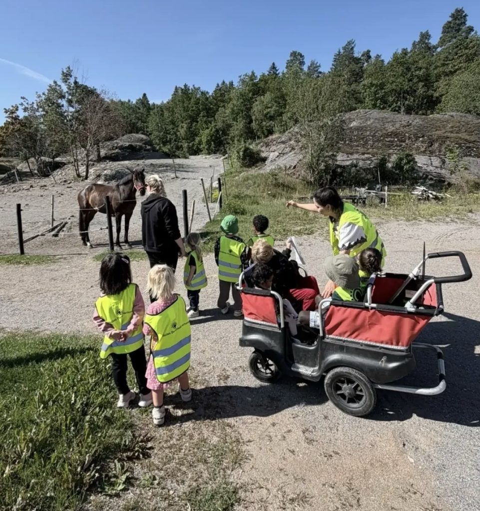 Förskolebarn i gula reflexvästar tittar på en brun häst innanför ett staket, medan några barn sitter i en fyrhjulig barnvagn med röd kapell. En vuxen pekar mot hästen. I bakgrunden syns klippor, träd och en klarblå himmel.