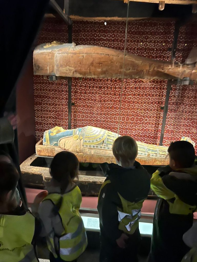 En gammal, dekorerad egyptisk mumiekista med en mumie i färgglada målningar visas i en monter på ett museum. Framför montern står flera barn i reflexvästar och tittar på föremålet.