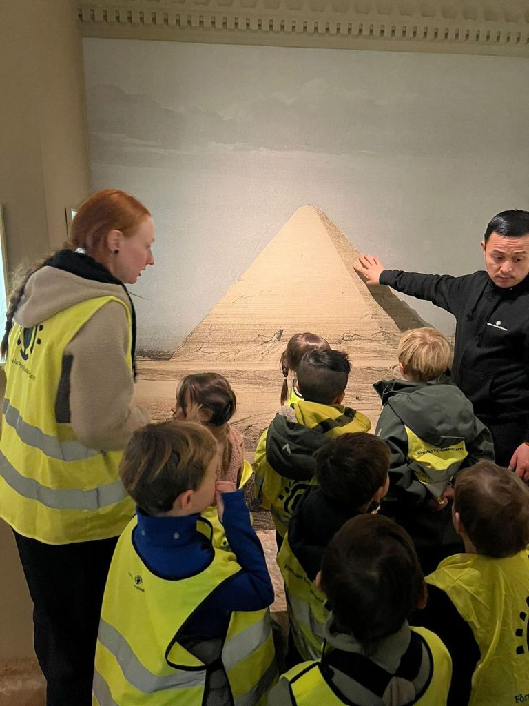 En grupp barn och en förskolepedagog med reflexvästar tittar på en museeguide som pekar på enegyptisk pyramid.