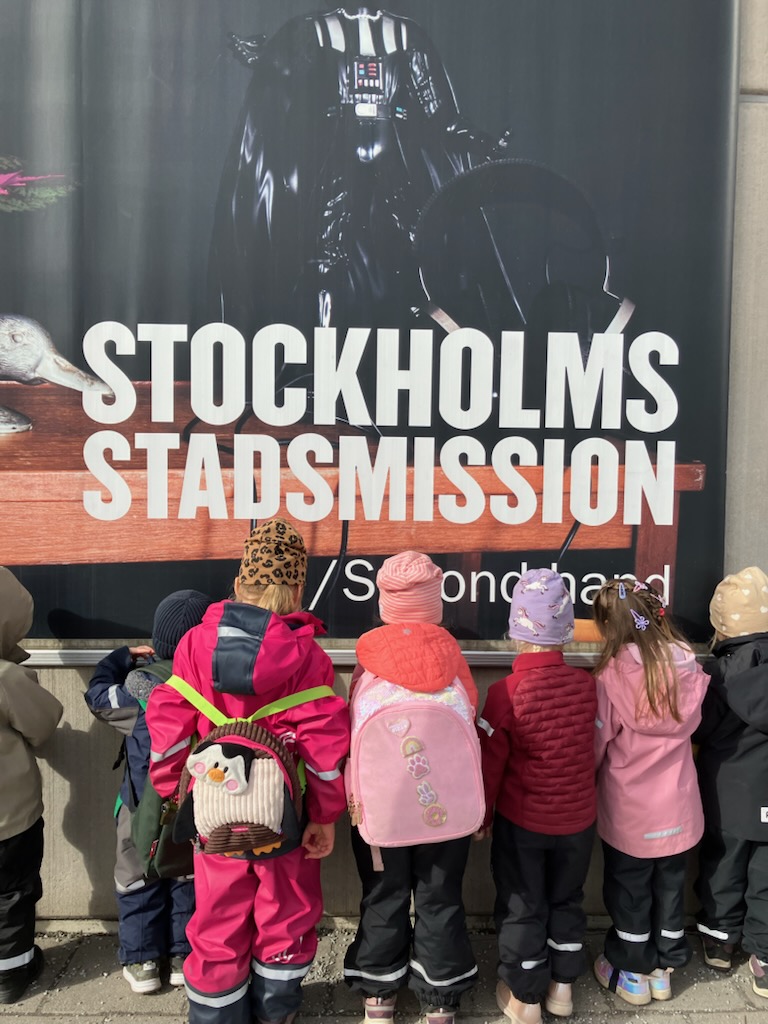 Några förskolebarn i färgglada overaller, jackor och ryggsäckar står uppradade med ryggen mot kameran framför en stor affisch med texten "Stockholms Stadsmission Secondhand". Över texten syns en mörk figur i hjälm och mantel.